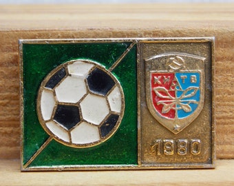Puede incluir: Un pin de color dorado con un campo de fútbol verde y dorado, un balón de fútbol y un escudo con un diseño rojo y azul. El escudo tiene el texto "KHTB" y el año "1980" está impreso debajo del escudo.