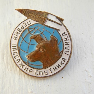 May include: Vintage metal pin featuring a brown dog's head inside a blue globe, with Cyrillic text and a rocket. The pin has a gold-colored border and a white and gold rocket design. The text reads "First Passenger Sputnik Laika".