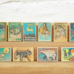 May include: Ten vintage matchboxes with colorful illustrations of animals and scenes. The matchboxes are arranged in two rows on a wooden surface. The top row features a crow, a man in a boat, a deer, a fox, a goat, and a ship. The bottom row features a fox, a monkey, a polar bear, and two figures in a forest.
