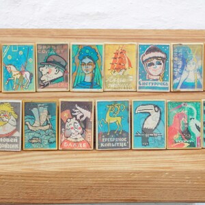 May include: A collection of 16 vintage matchboxes with colorful illustrations. The matchboxes feature various images, including a cartoon pig, a carousel horse, a toucan, a deer, and a woman in a blue dress. The matchboxes are arranged in two rows on a wooden surface.