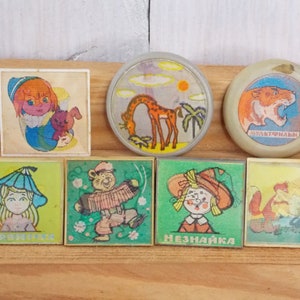May include: Six vintage, colorful, square and round pins with cartoon characters and animals. The pins feature a giraffe, a tiger, a girl with a blue hat, a girl with a red hat, a bear playing an accordion, and a girl with a red hat. The pins have Russian text on them.