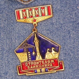 Puede incluir: Una medalla de color dorado con un diseño azul y dorado. La medalla tiene una cinta roja y blanca con el texto "XXXV" en ella. La medalla también tiene el texto "СТРОИТЕЛЯМ БАЙКОНУРА" en ella.