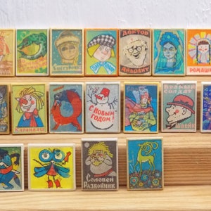 May include: A collection of 18 vintage matchboxes with colorful illustrations and Russian text. The matchboxes feature various characters and scenes, including a snowman, a doctor, a girl with flowers, and a wolf. The matchboxes are arranged in three rows on a wooden surface.
