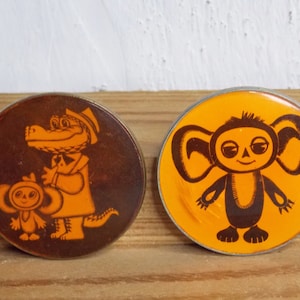 May include: Two round vintage pins. One pin features a brown and orange illustration of a crocodile and a small Cheburashka character. The other pin has an orange background with a Cheburashka character.