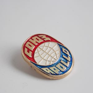May include: A vintage pin with a gold border, featuring the words "COHO" in red and "APOLLO" in blue. The center has a white globe design. The pin is oval-shaped and likely made of metal.