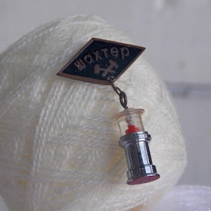 May include: A metal pin featuring a black diamond-shaped emblem with the Cyrillic text "шахтер" and a hammer and pick symbol. A small, dangling charm shaped like a vintage miner's lamp with a silver-toned base and a red top hangs from the emblem.