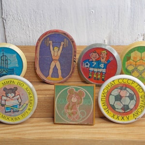 May include: Seven round, colorful, vintage pins with various sports themes. The pins feature images of weightlifting, ice hockey, soccer, and the Olympic rings. The pins are on a wooden surface.