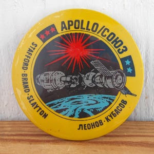 May include: A yellow round pin with the text "APOLLO/COHO3" and the names "STAFFORD - BRAND - SLAYTON" and "LEONOV - KUBASOV" around the edge. The pin features a red starburst and a black and white illustration of a spacecraft.