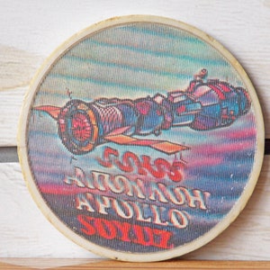 May include: A round, white, plastic button with a colorful image of a spacecraft in space. The text "SOYUZ APOLLO 75" is printed on the button.