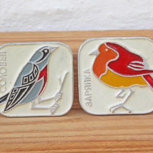 May include: Two vintage enamel pins featuring birds. The first pin is a blue and black bird with the word "СОЛОВЕЙ" written in white. The second pin is a red and yellow bird with the word "ЗАРЯНКА" written in white.