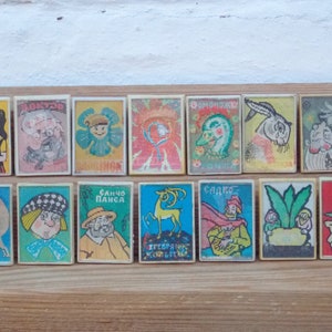 May include: A collection of 15 vintage matchboxes with colorful illustrations. The matchboxes feature various characters and scenes, including a woman in a red dress, a man with a beard, a flower, a deer, and a rabbit. The matchboxes are arranged in three rows of five.