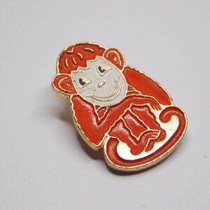 Pode incluir: Um alfinete de macaco vintage com uma borda dourada. O macaco é retratado em esmalte vermelho com um rosto branco e uma expressão sorridente. Este alfinete colecionável é um acessório divertido.