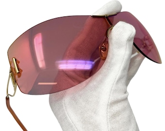 Puede incluir: Un par de gafas de sol rosas con montura dorada. Las gafas de sol tienen un diseño sin montura y se sostienen en un paño blanco.
