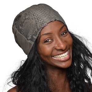 May include: A woman with long, dark hair is wearing a grey beanie with a snakeskin pattern on the top. She is smiling and looking to the side.