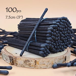 Puede incluir: Un paquete de 100 cerillas de madera negra, cada una de 7,5 cm de largo, con cabezas de color gris oscuro. Las cerillas están atadas con un hilo azul fino y descansan sobre una rodaja de madera circular, rodeadas de cerillas sueltas.