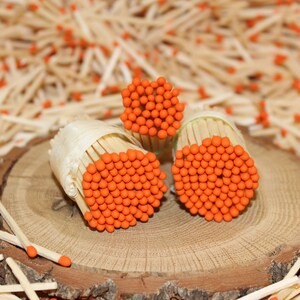 May include: Three bundles of matches with orange heads are arranged on a wooden surface. The matches are wrapped in white paper and are surrounded by loose matches.