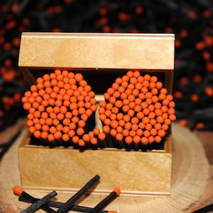 May include: A wooden matchbox containing two clusters of orange-headed matches. The matchsticks are black, and the box is made of light-coloured wood. Several loose matches are scattered in front of the box, with a dark background.