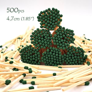 May include: A collection of wooden matches with green tips, some bundled together. The image shows approximately 500 matches, with the text indicating a length of 1.85 inches (4.7 cm). The matches are scattered on a white surface.