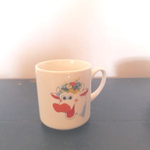 May include: A white ceramic mug featuring a colorful graphic of a cow's head wearing a crown of small flowers. The cow has a bright red nose, red inner ears, and a wide, smiling mouth. The mug has a simple cylindrical shape with a curved handle.