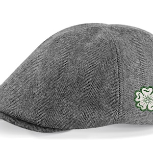 Puede incluir: Una gorra plana de color gris con estampado de espiga y un trébol verde bordado en el lateral.