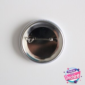 Op de afbeelding: De achterkant van een ronde knop met een zilveren pin en een witte achtergrond. De knop heeft een blauwe en witte rand. De tekst op de knop luidt "Cadeau qui D&eacute;chire" met sterren rond de tekst.