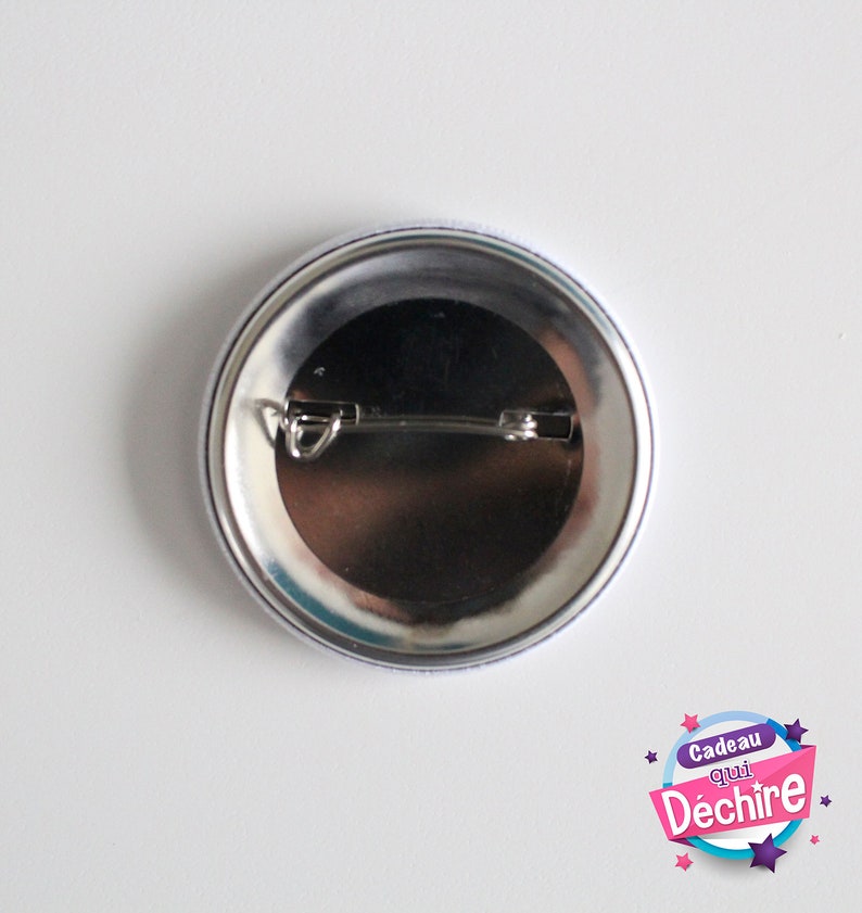Op de afbeelding: De achterkant van een ronde knop met een zilveren pin en een witte achtergrond. De knop heeft een blauwe en witte rand. De tekst op de knop luidt "Cadeau qui D&eacute;chire" met sterren rond de tekst.
