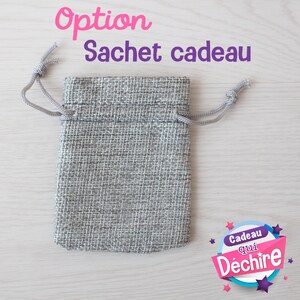 Op de afbeelding: Een grijze jute trekkoordtas met een wit koord. De tas ligt op een licht houten oppervlak. De tekst "Option Sachet cadeau" staat boven de tas. De tekst "Cadeau qui D&eacute;chire" staat in een cirkel met sterren en roze, blauwe en witte kleuren.