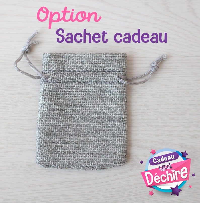 Op de afbeelding: Een grijze jute trekkoordtas met een wit koord. De tas ligt op een licht houten oppervlak. De tekst "Option Sachet cadeau" staat boven de tas. De tekst "Cadeau qui D&eacute;chire" staat in een cirkel met sterren en roze, blauwe en witte kleuren.