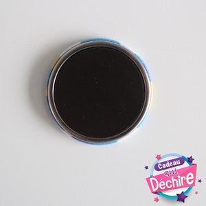 Op de afbeelding: Een ronde magneet met een zwart midden en een kleurrijke rand. De rand heeft een blauw, geel en wit patroon. De magneet heeft de tekst "Cadeau qui D&eacute;chire" erop.