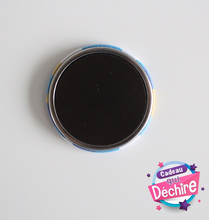 Op de afbeelding: Een ronde magneet met een zwart midden en een kleurrijke rand. De rand heeft een blauw, geel en wit patroon. De magneet heeft de tekst "Cadeau qui D&eacute;chire" erop.