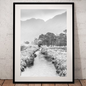 May include: A black and white photograph of a stream flowing through a misty valley with trees and mountains in the background.