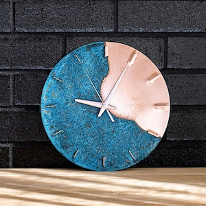 A copper wall clock with a vibrant, blue patina sits atop a wooden bench and resting against a black, brick wall. The clock has copper hands. The clock is made by the company Copper Brass and Bronze and Empire Copper by  Hayes Home.