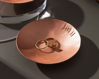 May include: Two gold rings, one with a small gemstone, are displayed on a copper-colored dish with a textured surface. The dish has a series of lines etched into the surface.