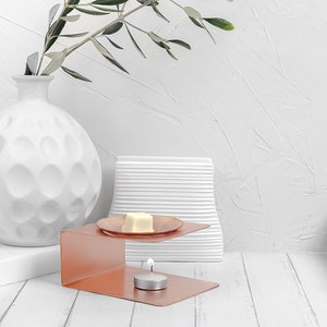 May include: A copper-colored metal diffuser with a white wax melt on a small plate. The diffuser is on a white wooden surface with a white ceramic vase and a white ceramic object behind it.