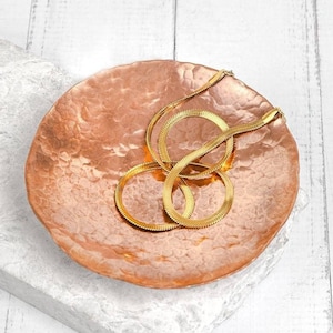Könnte beinhalten: Eine goldene Schlangenkettenkette liegt auf einer gehämmerten Kupferschale. Die Schale befindet sich auf einer weißen Oberfläche.