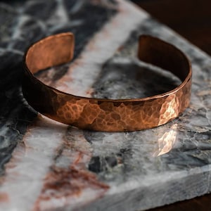 May include: A hammered copper bracelet with a smooth, polished finish. The bracelet is lying on a marble surface.