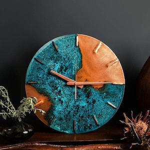 A copper wall clock with a vibrant, blue patina sits atop a wooden bench and in front of navy a background. The clock is made by the company Copper Brass and Bronze and Empire Copper by  Hayes Home.