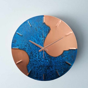 A copper wall clock with a stunning, electric, blue patina is photographed against a grey backdrop. The clock has copper hands and is made by the company Copper Brass and Bronze and Empire Copper by Hayes Home.