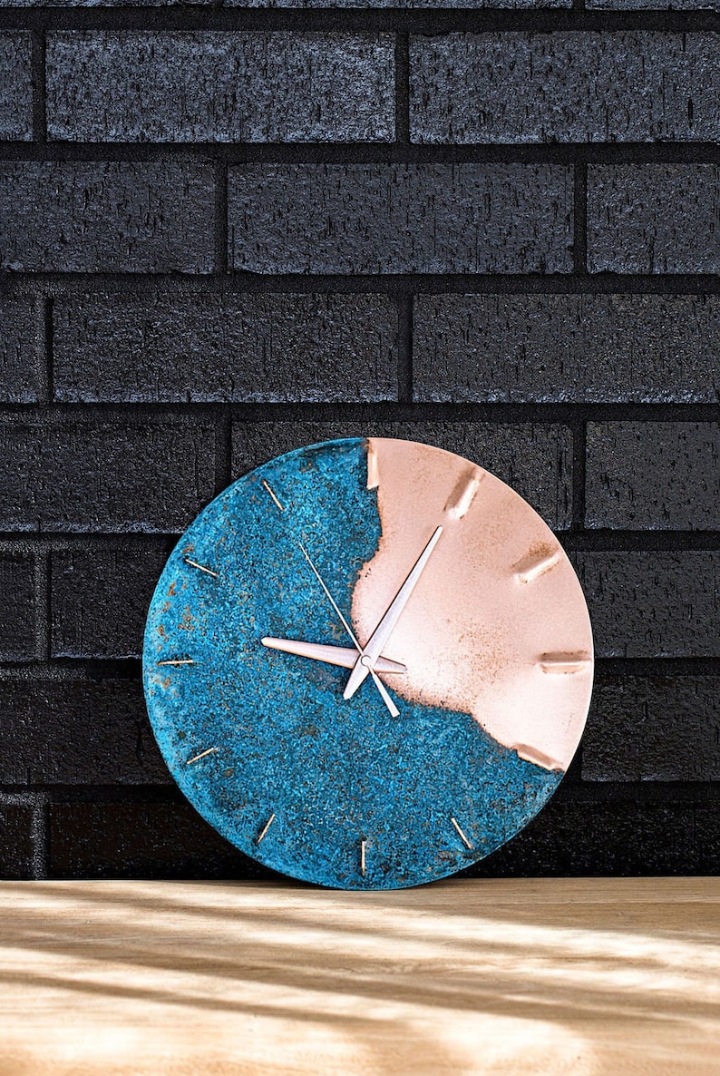 A copper wall clock with a vibrant, blue patina sits atop a wooden bench and resting against a black, brick wall. The clock has copper hands. The clock is made by the company Copper Brass and Bronze and Empire Copper by  Hayes Home.