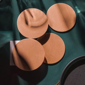 May include: Four round, light brown coasters on a dark green surface. The coasters are made of a textured material and have a subtle sheen.