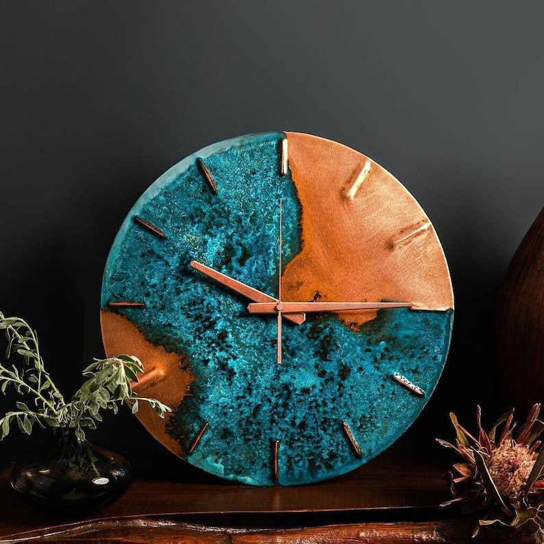 A copper wall clock with a vibrant, blue patina sits atop a wooden bench and in front of navy a background. The clock is made by the company Copper Brass and Bronze and Empire Copper by  Hayes Home.