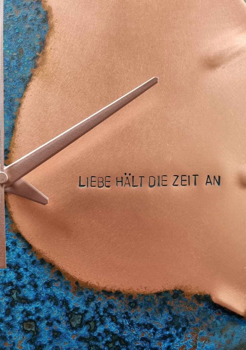 A close up image of the Blue Patina Copper Wall Clock with the engraving LIEBE HALT DIE ZEIT AN which translates to Love Stops Time. The clock is made by Copper, Brass and Bronze and Empire Copper by Hayes Home.