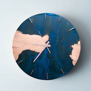 A copper wall clock with a vibrant, blue patina is photographed against a grey backdrop. The clock has copper hands and is made by the company Copper Brass and Bronze and Empire Copper by Hayes Home.