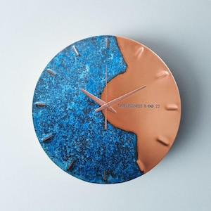 A copper wall clock with a vibrant, blue patina is photographed against a grey backdrop. The clock has copper hands and is engraved TIMELESSNESS 11 22. The clock is made by the company Copper Brass and Bronze and Empire Copper by Hayes Home.