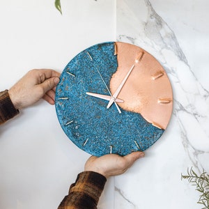 A copper wall clock with a vibrant, blue patina is held against a white, marble wall. The clock has copper hands and is made by the company Copper Brass and Bronze and Empire Copper by Hayes Home.