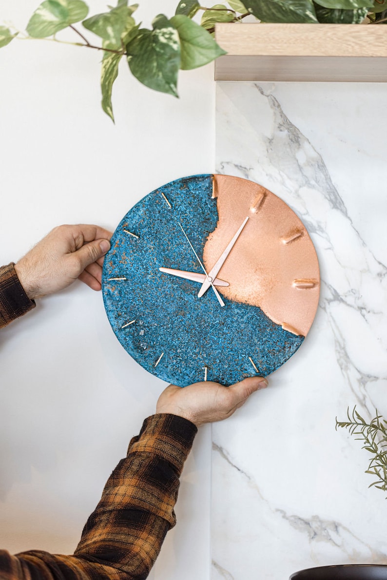 A copper wall clock with a vibrant, blue patina is held against a white, marble wall. The clock has copper hands and is made by the company Copper Brass and Bronze and Empire Copper by Hayes Home.