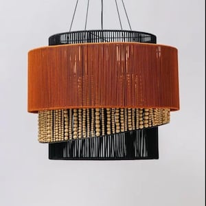 May include: A multi-layered pendant light. The top is black, followed by a burnt orange shade, a layer of wooden beads, and a black base. The light hangs from thin black wires, creating a modern design.