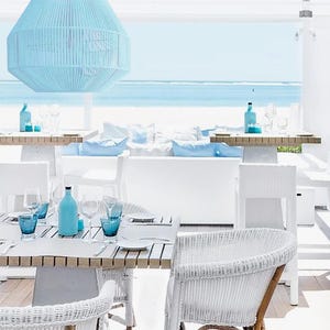 May include: Outdoor dining scene with a light blue pendant lamp, white furniture, and a view of the ocean. The table is set with blue glassware and bottles. White wicker chairs with wooden legs and accents surround the table.