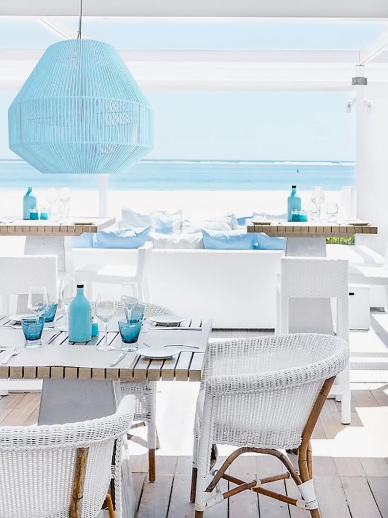 May include: Outdoor dining scene with a light blue pendant lamp, white furniture, and a view of the ocean. The table is set with blue glassware and bottles. White wicker chairs with wooden legs and accents surround the table.