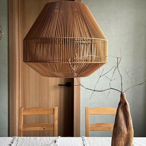 May include: A large, woven pendant light fixture hangs above a table. The light is made of tan-colored rope and has a geometric shape. A wooden vase with branches and a decorative wooden pod are on the table. Two wooden chairs are visible.